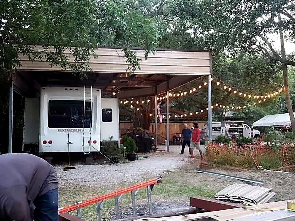 Completed steel RV cover with a tall clearance roof and festoon lighting over a gravel pad