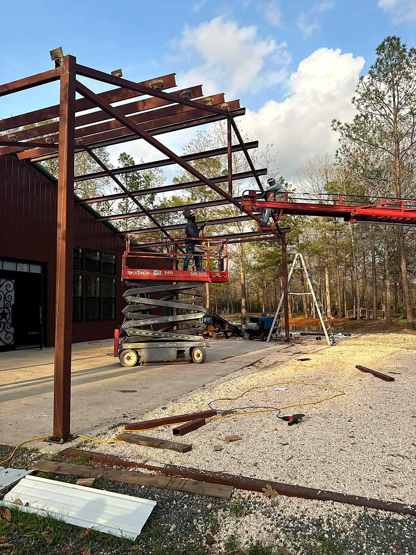 Crew welding a steel patio frame from scissor lifts beside a metal building